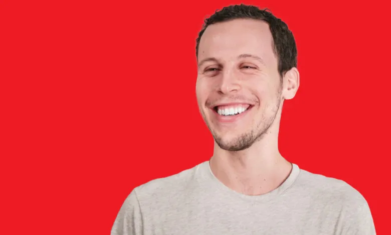 A man with short dark hair and a beard smiles broadly against a solid red background.