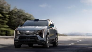 A gray Cadillac Lyriq electric SUV drives on a highway with trees and mountains in the background.