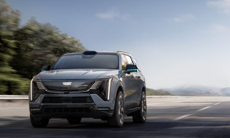 A gray Cadillac Lyriq electric SUV drives on a highway with trees and mountains in the background.