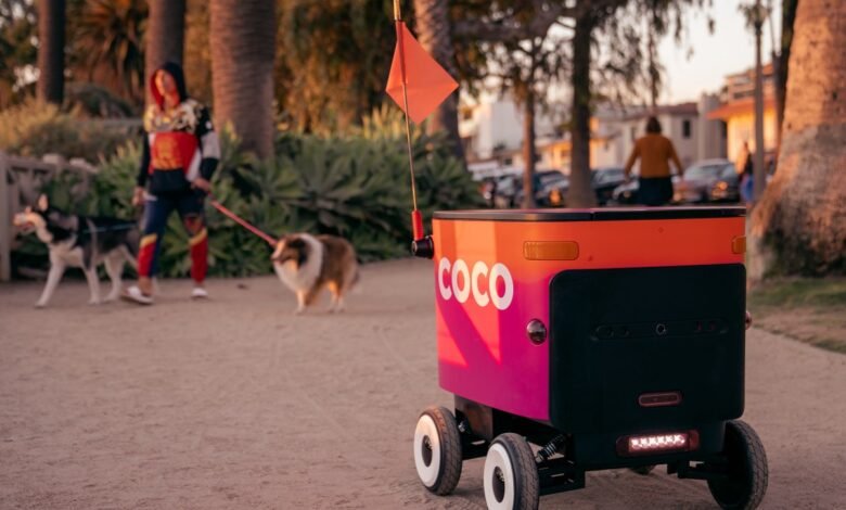 A pink and orange delivery robot with the 'COCO' logo on a path, with two dogs and a person walking in the background.