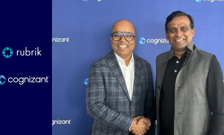 Two men smiling and shaking hands in front of a blue backdrop with Cognizant and Rubrik logos.