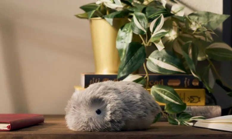 A fluffy grey robotic pet sits on a wooden desk next to books and a plant.