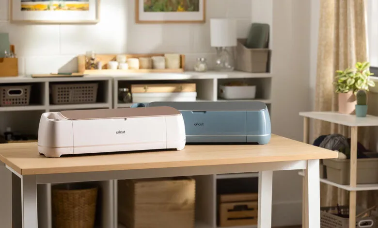 Two Cricut Maker cutting machines, one in seashell pink and one in sage green, on a wooden table.