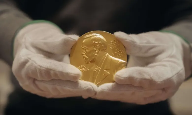 Hands wearing white gloves hold a gold Nobel Prize medal featuring Alfred Nobel's profile.