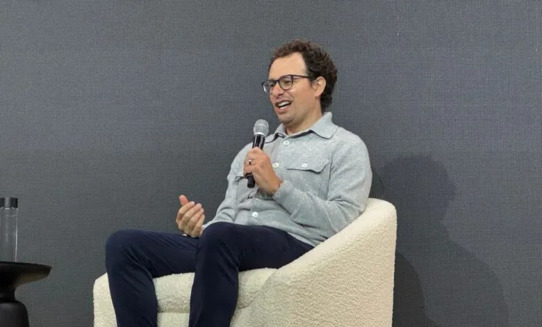 Man with curly hair and glasses speaking into a microphone while seated.