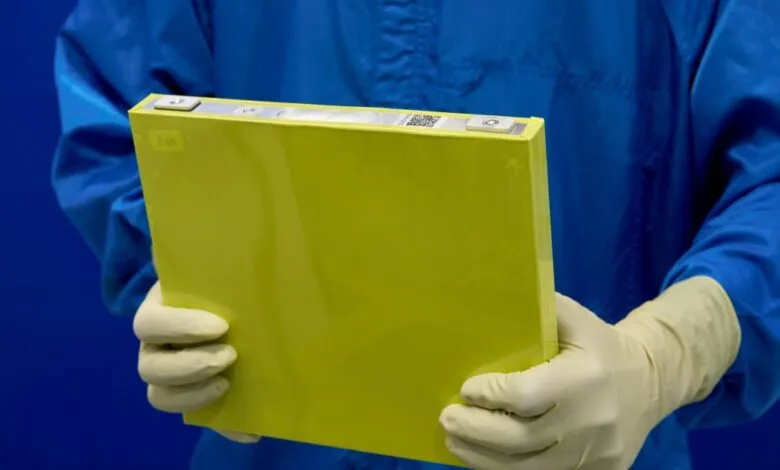 Person in blue lab coat and gloves holding a bright yellow battery cell.