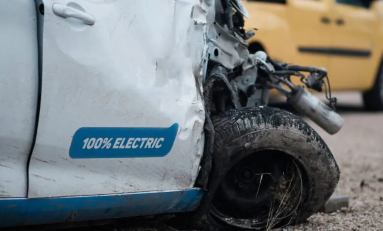 A severely damaged white electric car with '100% ELECTRIC' visible on the side.