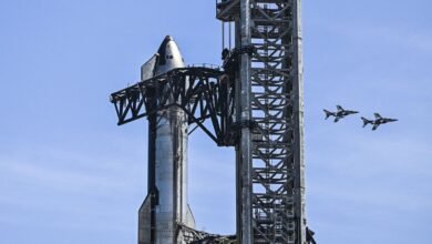 A large, silver rocket stands next to a towering metal launch structure, with two fighter jets flying in formation against a clear blue sky.