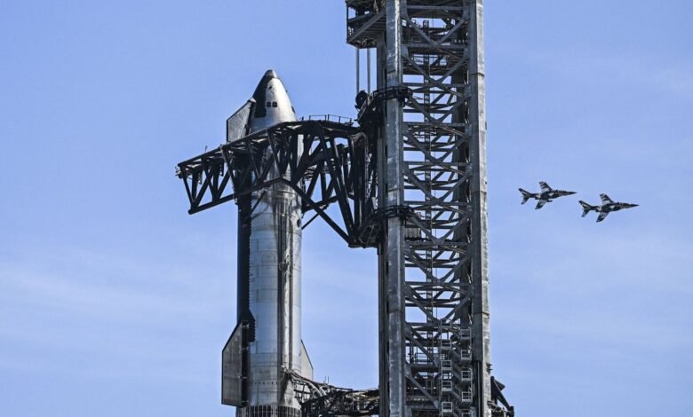 A large, silver rocket stands next to a towering metal launch structure, with two fighter jets flying in formation against a clear blue sky.