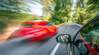View from inside a car as a red car overtakes on a tree-lined road with motion blur.