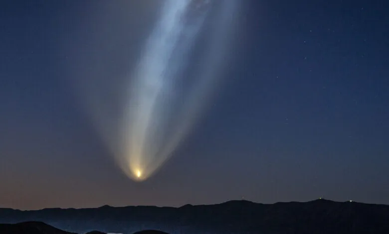 A SpaceX Falcon 9 rocket ascends into the dark blue night sky, leaving a bright, glowing plume of exhaust.