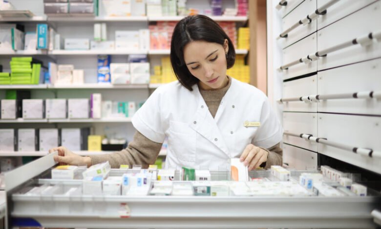 A female pharmacist in a white coat organizes medication in a pharmacy storage drawer.