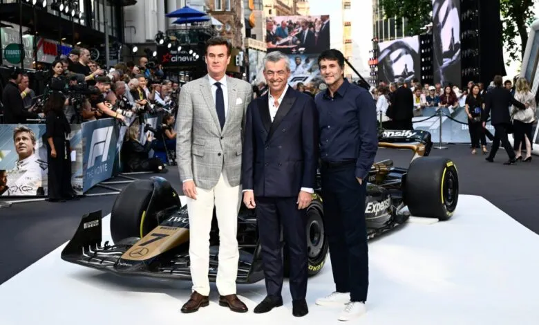 Three men in suits stand next to a Formula 1 race car on a red carpet.