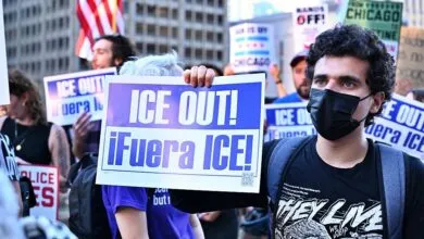 Man in black mask holds sign reading 'ICE OUT! ¡Fuera ICE!' at a protest.