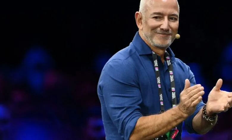 Jeff Bezos speaking on stage with a microphone, wearing a blue shirt and a lanyard.