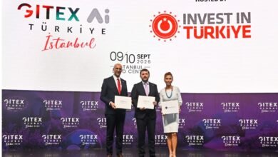 Three people in formal attire holding certificates on a stage with event branding.