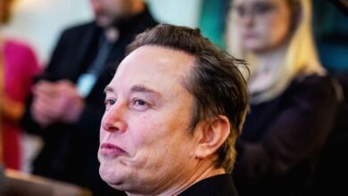 Close-up profile of Elon Musk looking intently to the left, with blurred figures in the background.