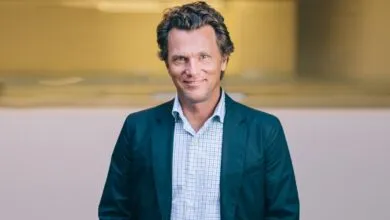 A smiling man in a suit jacket and checkered shirt looks directly at the camera.