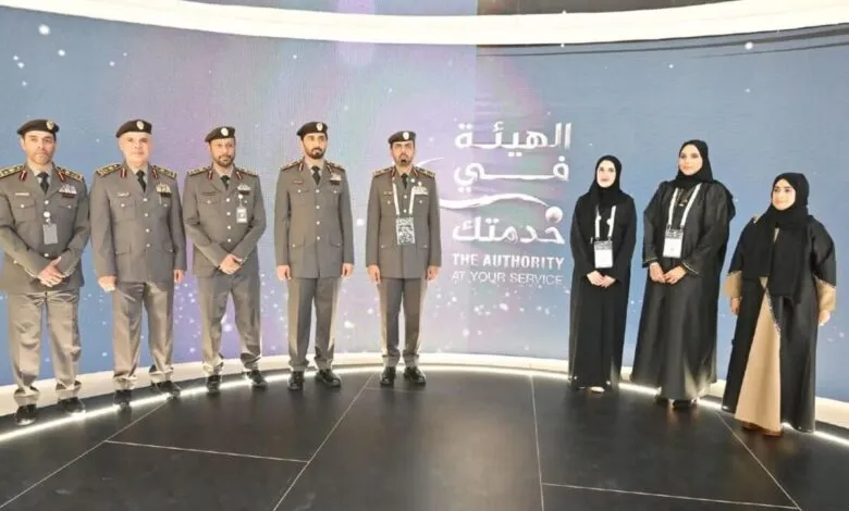 Group of uniformed men and women stand in front of a screen with Arabic and English text.