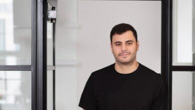 A man in a black t-shirt stands in an office doorway with glass partitions.