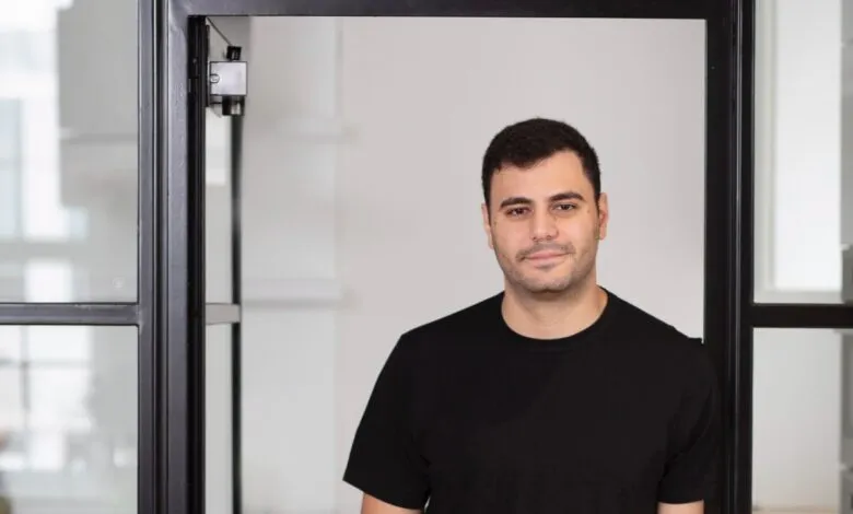 A man in a black t-shirt stands in an office doorway with glass partitions.