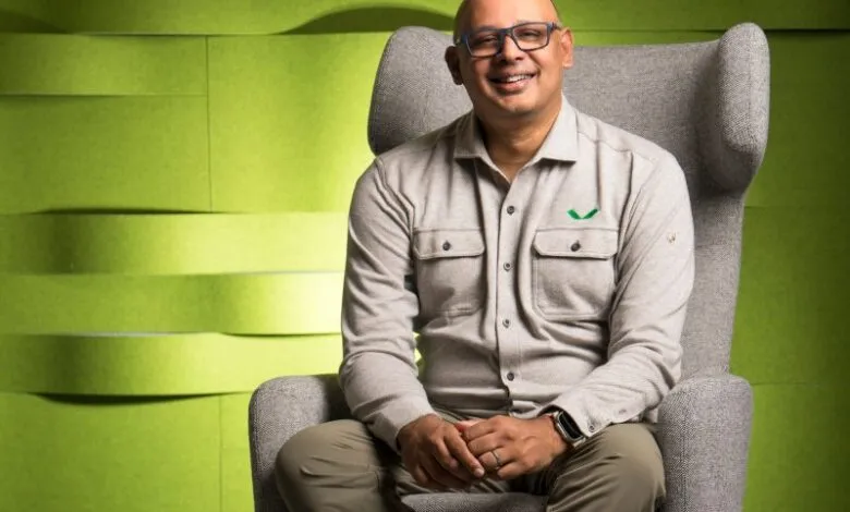 Man in a grey shirt and olive pants sitting in a grey armchair, smiling.