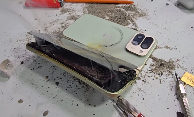 A damaged light green smartphone with its screen peeled back, showing smoke and debris.