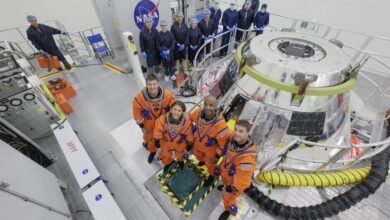 Astronauts in orange suits pose with a spacecraft module and NASA personnel in a cleanroom.