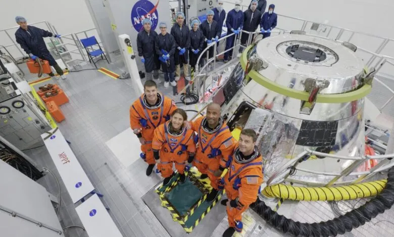 Astronauts in orange suits pose with a spacecraft module and NASA personnel in a cleanroom.