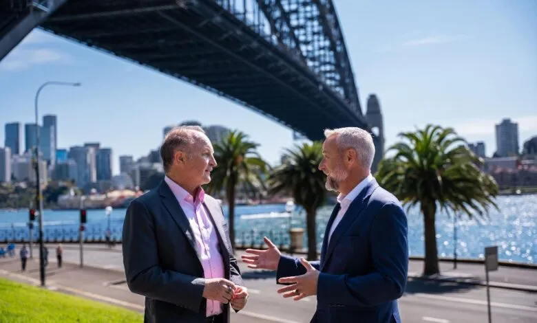 Two businessmen in suits converse outdoors with Sydney Harbour Bridge and skyline in background.