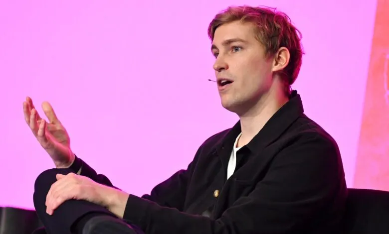 Man with blond hair speaking with his hand raised, against a pink background.