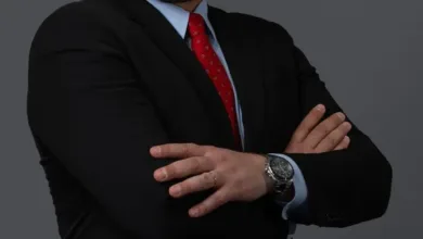 Bald man with a beard wearing a black suit, blue shirt, and red tie, arms crossed.