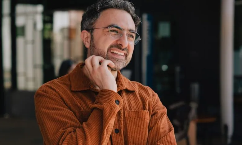 Man with graying hair and beard wearing glasses and an orange corduroy shirt, smiling.