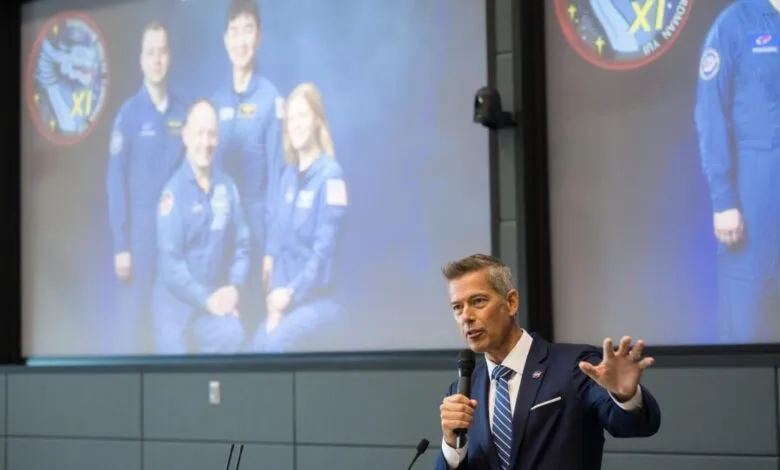 Man in suit speaking into microphone with astronauts projected on screen behind him.