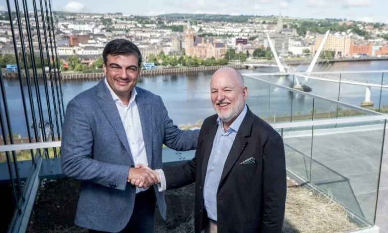 Two men in suits shake hands on a balcony overlooking a city and river.