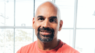 Bald man with a beard wearing a coral t-shirt smiles warmly in front of a large window.