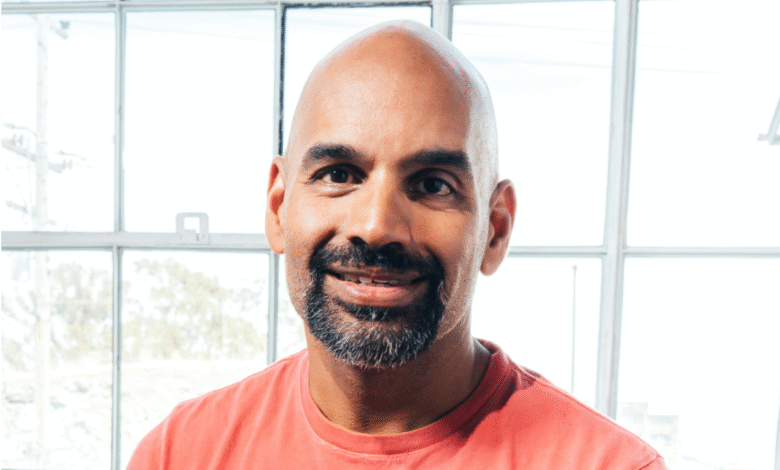 Bald man with a beard wearing a coral t-shirt smiles warmly in front of a large window.