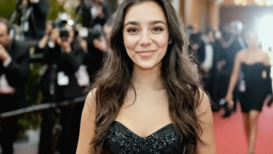 Young woman in a black dress smiles on a red carpet with photographers in the background.