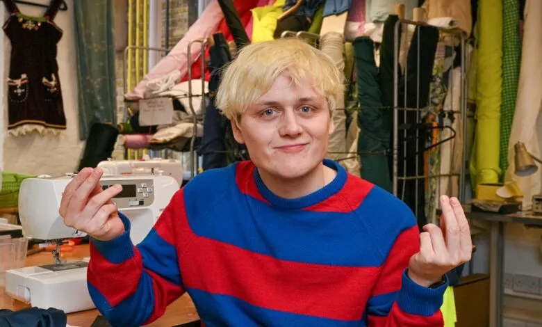 A young person with blonde hair and blue eyes smiles, gesturing with their hands in a workshop.