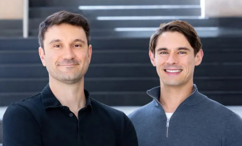 Two smiling men in business casual attire stand in front of a modern staircase.