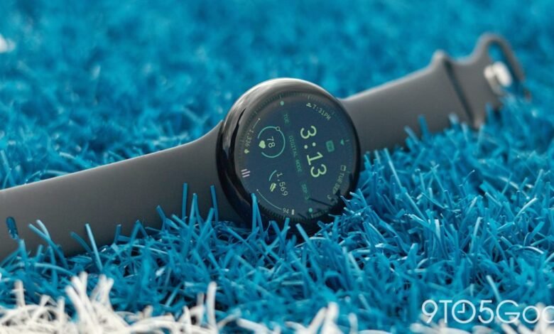 Close-up of a dark grey smartwatch with a blue watch face on bright blue artificial turf.