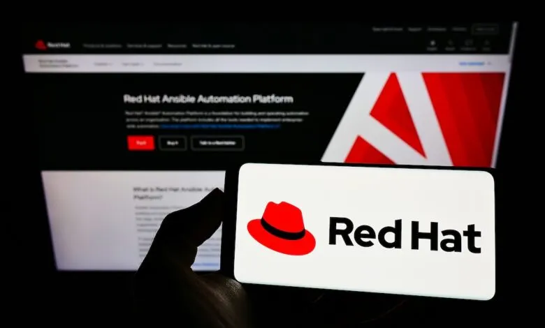 A hand holds a smartphone displaying the Red Hat logo and name, with a computer screen in the background showing Red Hat Ansible Automation Platform.