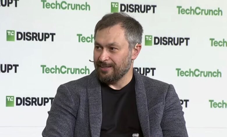 Man with beard and grey jacket speaking on stage at TechCrunch Disrupt event.