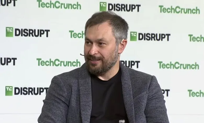 Man with beard and grey jacket speaking on stage at TechCrunch Disrupt event.