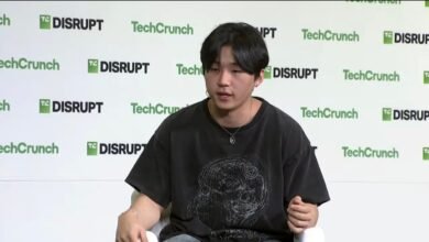 Young man with dark hair wearing a graphic t-shirt and necklace, speaking at an event.