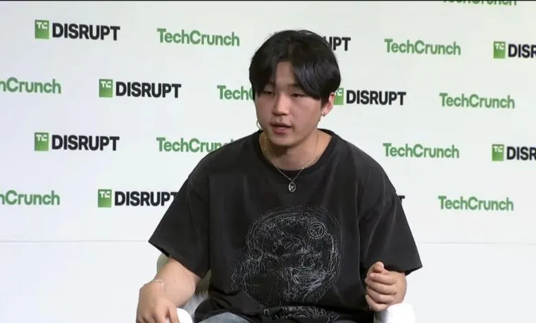 Young man with dark hair wearing a graphic t-shirt and necklace, speaking at an event.