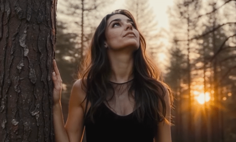 Woman in black dress looking up at the sky, leaning against a tree in a forest at sunset.