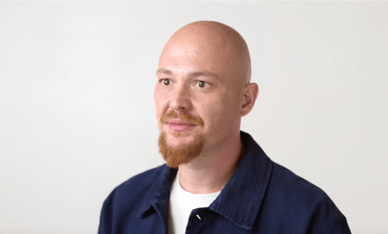 Bald man with a goatee wearing a dark blue shirt and white t-shirt, looking to the side.
