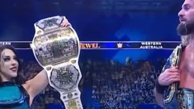 A woman holds up a championship belt while a man stands beside her, both wearing championship belts.