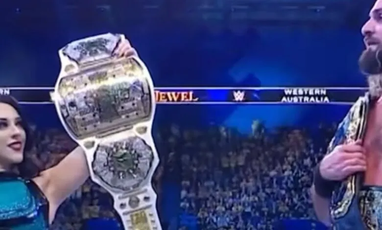 A woman holds up a championship belt while a man stands beside her, both wearing championship belts.